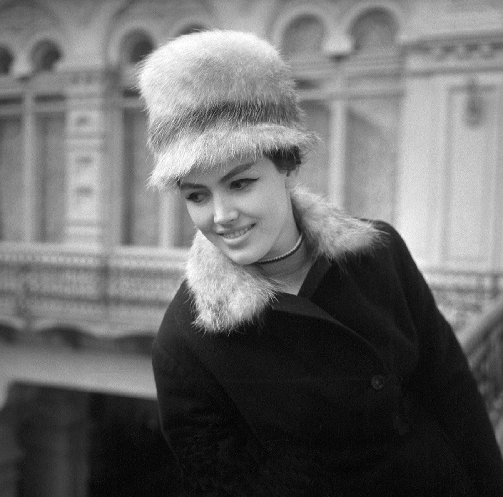 A young woman in a tall fur hat and a fur collared dark coat smiles on a gallery inside the GUM department store in Moscow, the arcaded galleries out of focus behind her