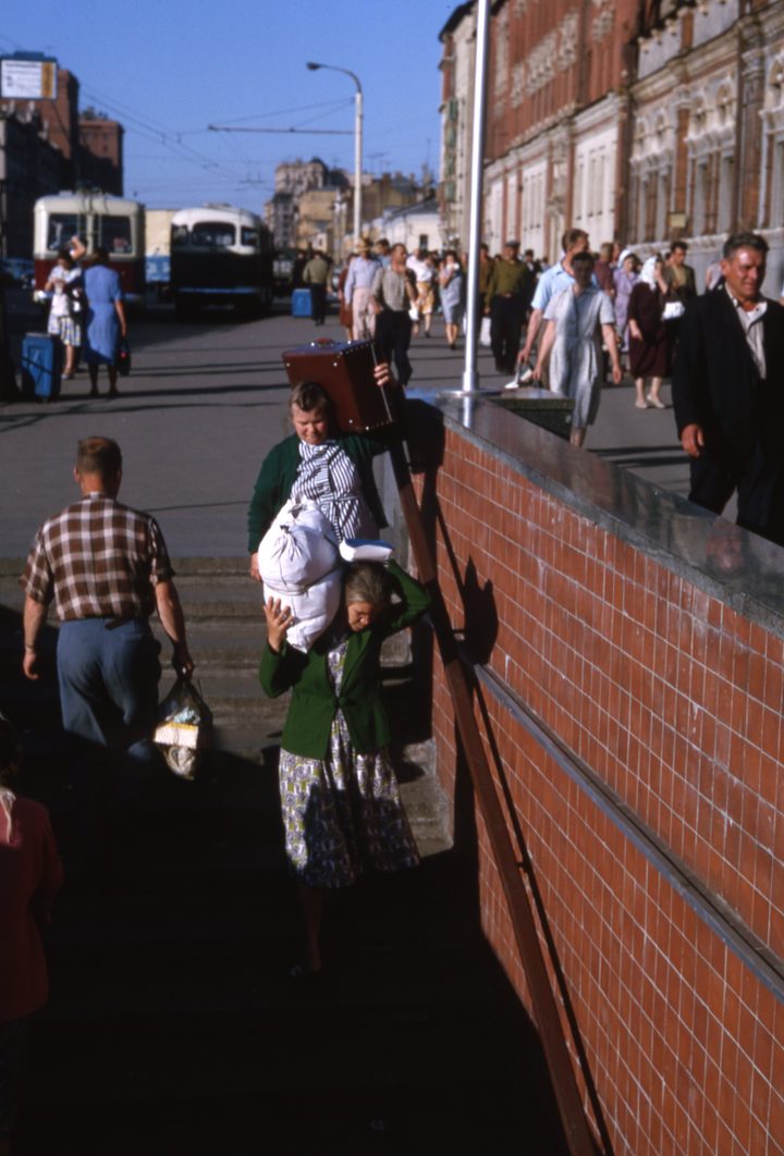 A colour slide of a Moscow railway square: travellers carrying suitcases and bundles down the steps of a pedestrian subway beside the red brick flank of the terminal, crowds on the pavement and trolleybuses on the street behind