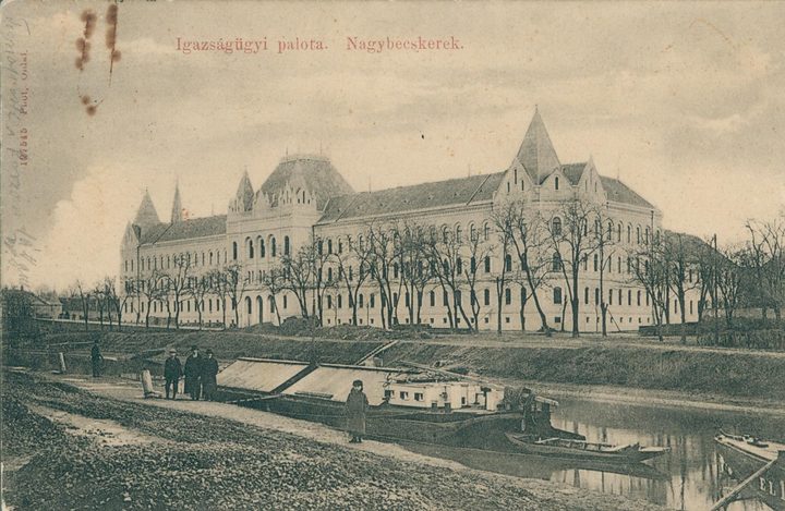 A period postcard captioned Igazsagugyi palota, Nagybecskerek: a long courthouse with corner turrets and rows of arched windows seen across a canal, barges moored at the near bank and four men standing on the towpath
