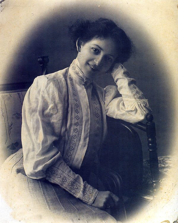 A studio photograph of a woman in an embroidered white blouse, seated sideways on a chair with her head tilted against her raised hand, smiling towards the camera