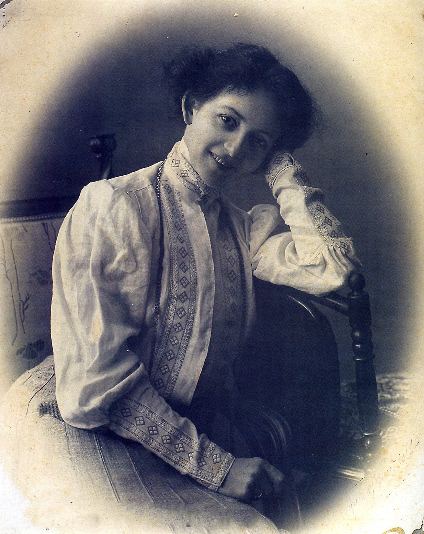 A studio photograph of a woman in an embroidered white blouse, seated sideways on a chair with her head tilted against her raised hand, smiling towards the camera