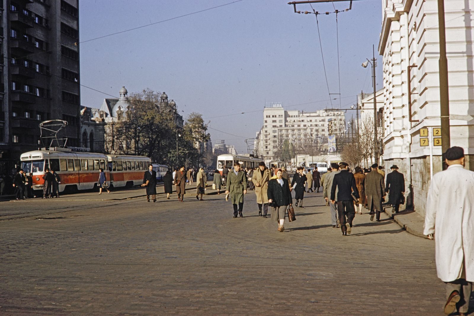 A colour photograph of a Bucharest street in 1970: people in overcoats walking along a wide cobbled pavement, a red and cream tram at a stop on the left and a rooftop sign reading Alba in the distance