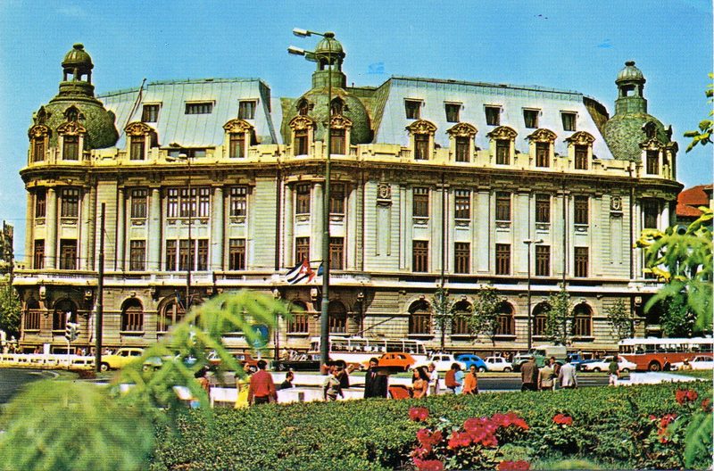 The University of Bucharest building in the 1970s, buses and cars passing on the boulevard in front of it and a flowerbed in the foreground