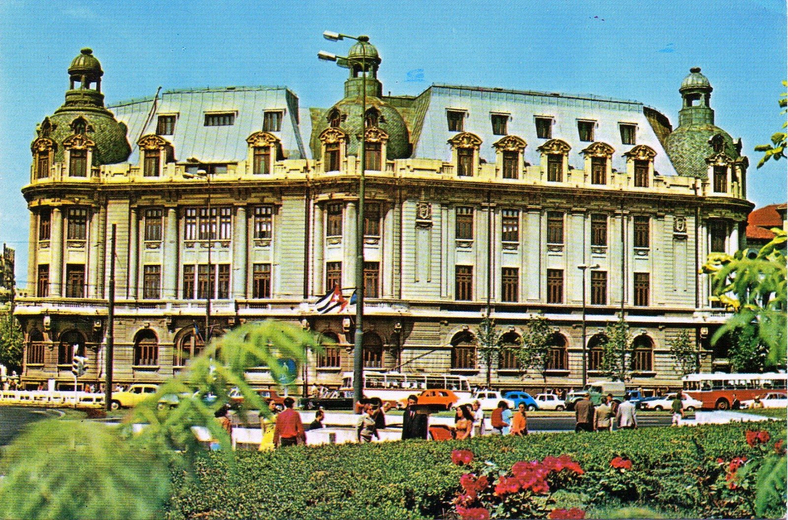 The University of Bucharest building in the 1970s, buses and cars passing on the boulevard in front of it and a flowerbed in the foreground