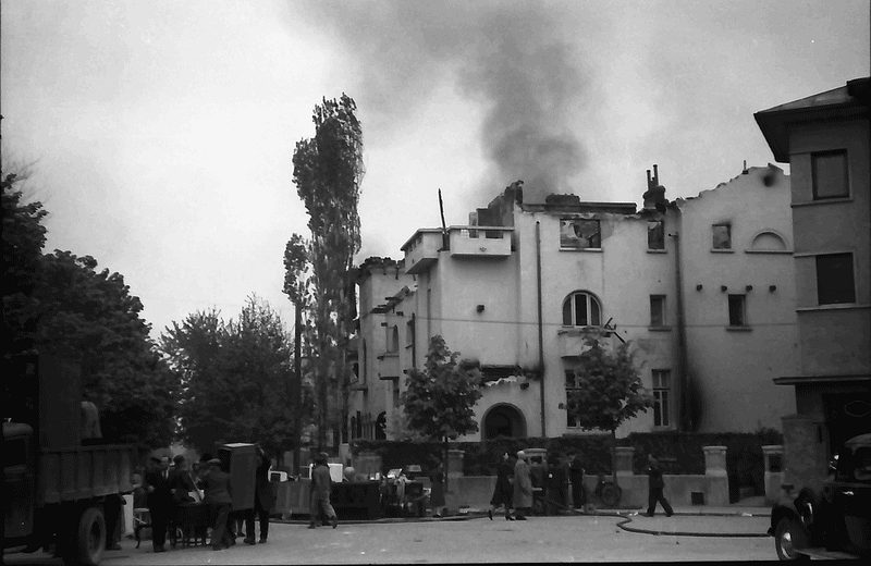 A large Bucharest house burning after an air raid in 1944, smoke pouring from the roof, people and a fire hose in the street in front of it