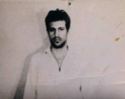A faded full length photograph of Ion Rîmaru standing against a plain wall in a light shirt and dark trousers, his hands at his sides