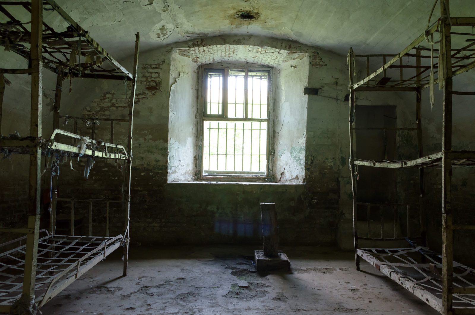 An abandoned vaulted cell in Fort 13 Jilava, rusted iron bunk frames along both walls, peeling plaster and a small barred window at the far end