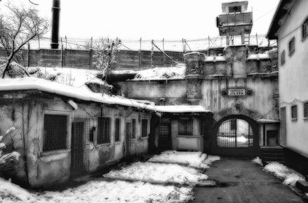 A snow covered yard inside Jilava prison, low cell buildings with barred windows, a wall topped with barbed wire and a guard tower above the gate