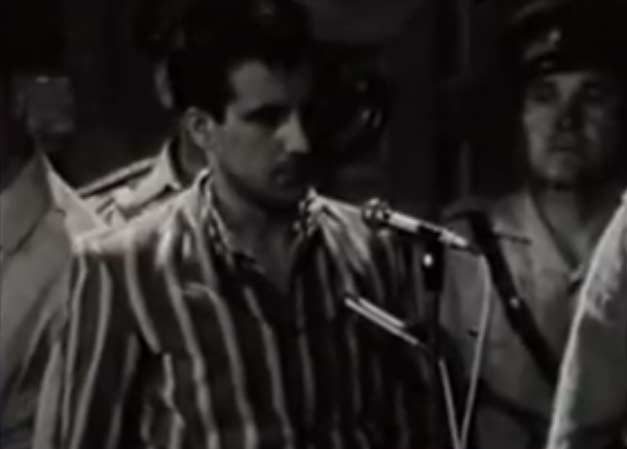 A frame from the 1971 trial broadcast: Ion Rîmaru in a striped prison shirt, head lowered at a standing microphone, with a uniformed Militia officer in a peaked cap beside him