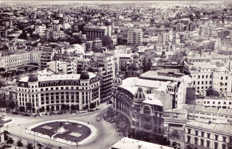 A black and white view over Bucharest from above in the early 1960s, University Square and its traffic circle in the foreground and a rooftop slogan in Romanian reading Traiasca Republica Populara Romina, long live the Romanian People's Republic