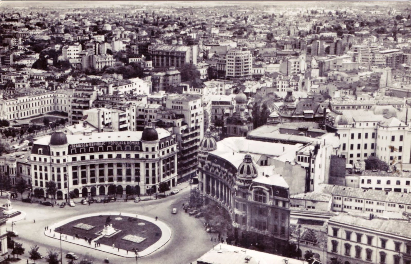 A black and white view over Bucharest from above in the early 1960s, University Square and its traffic circle in the foreground and a rooftop slogan in Romanian reading Traiasca Republica Populara Romina, long live the Romanian People's Republic