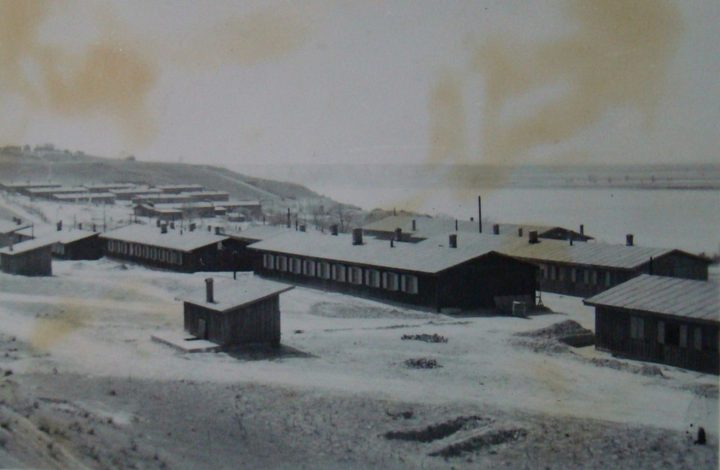 Rows of long wooden barracks on bare open ground beside a wide waterway, photographed from a rise, with further camp buildings in the distance