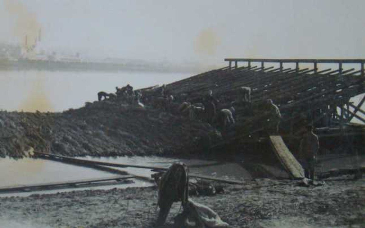 Men working by hand on a timber and earth embankment running into the water, a guard standing at the near end, industrial buildings on the far bank