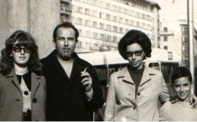 A black and white street photograph of four people on a Bucharest pavement in the 1970s: a man in a dark coat with a cigarette, two women in glasses and a child, apartment blocks and a bus behind them