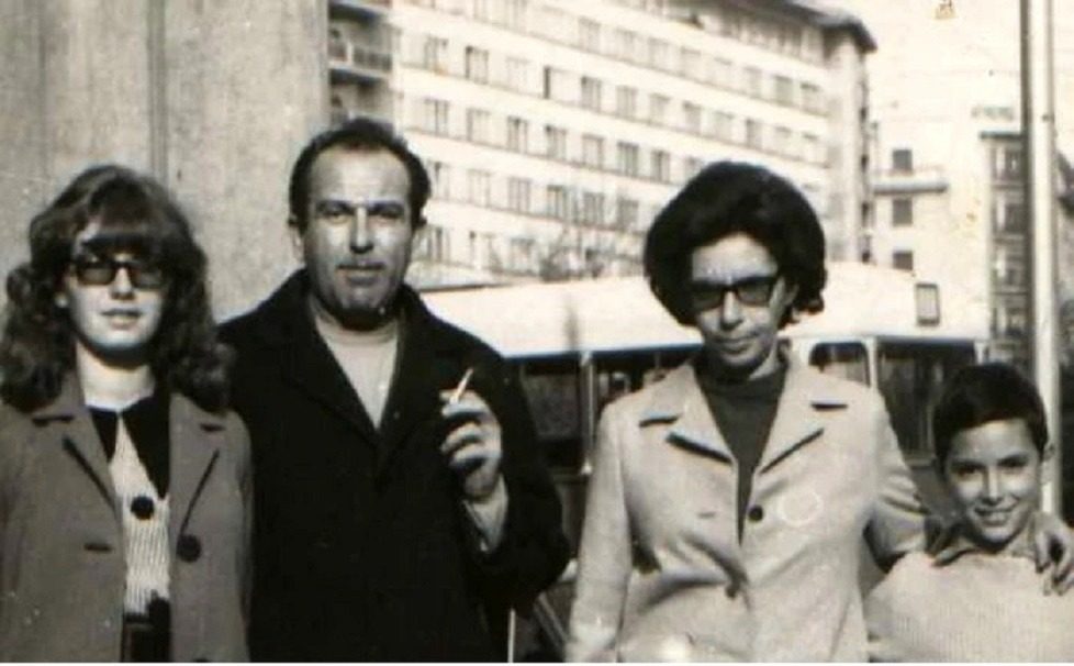 A black and white street photograph of four people on a Bucharest pavement in the 1970s: a man in a dark coat with a cigarette, two women in glasses and a child, apartment blocks and a bus behind them