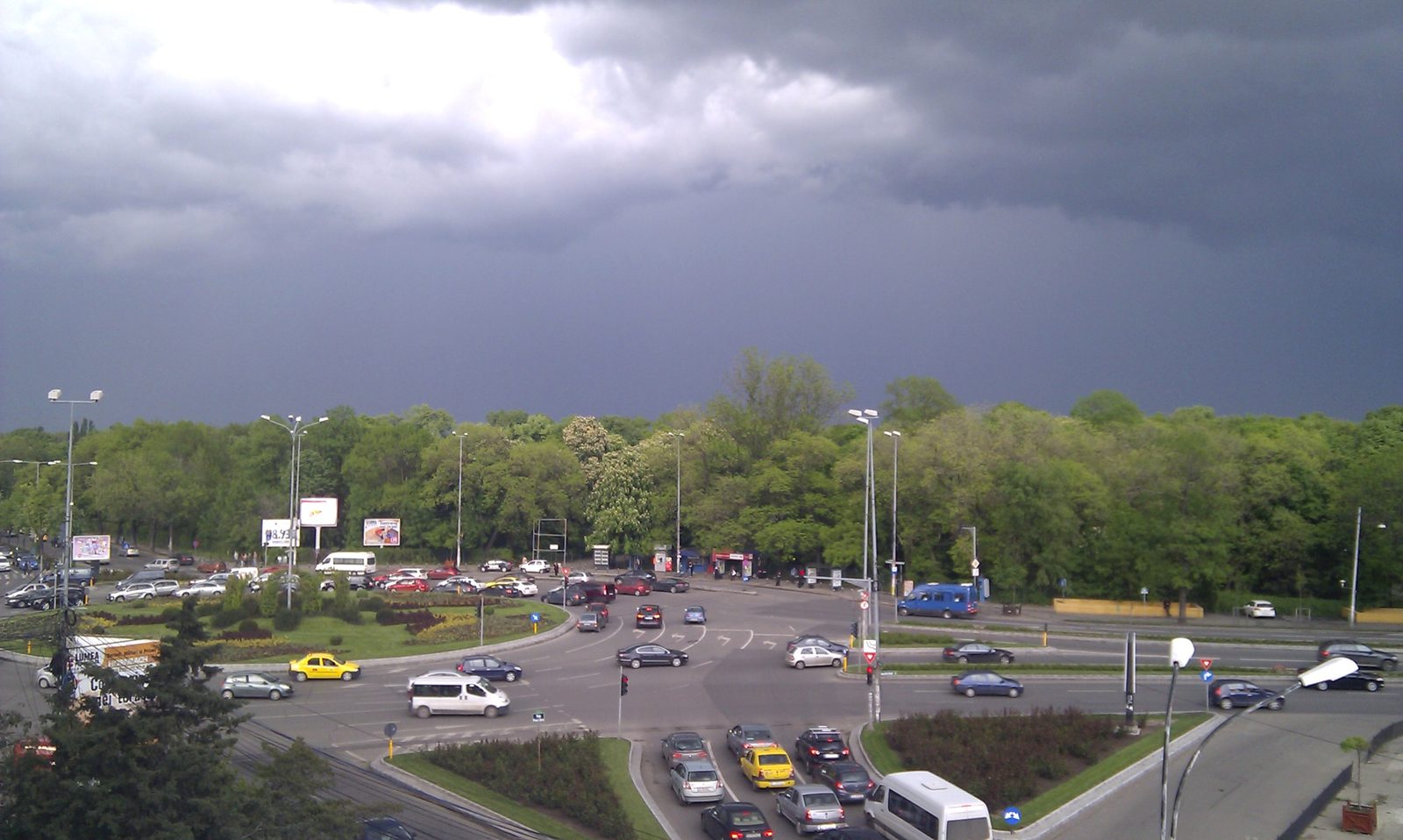 The edge of Herastrau Park in Bucharest seen from above under a storm sky: a boulevard junction with traffic in the foreground and the park's dense tree line running the width of the picture behind it
