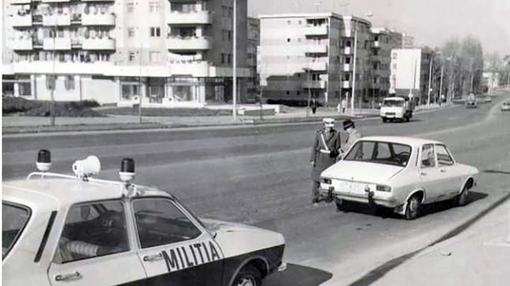 A black and white street photograph: a patrol car lettered MILITIA in the foreground and two militiamen stopping a white Dacia saloon across the road, apartment blocks behind