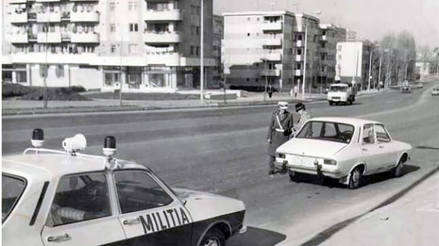 A black and white street photograph: a patrol car lettered MILITIA in the foreground and two militiamen stopping a white Dacia saloon across the road, apartment blocks behind