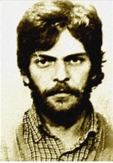 Head and shoulders police photograph of a young bearded man with long dark hair in a checked shirt, looking straight at the camera