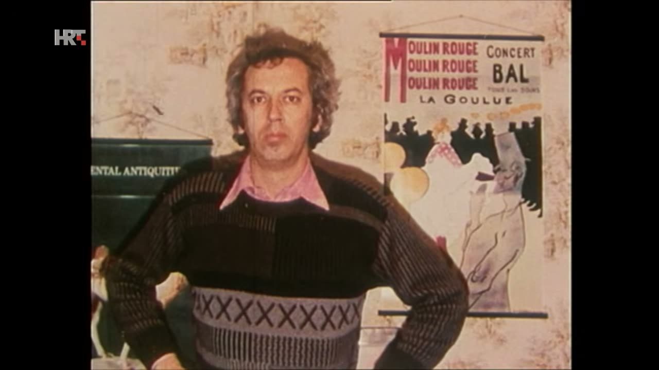 A colour film frame of Bruno Busic, a man with dark curly hair in a patterned jumper, standing with his hands on his hips in front of a Moulin Rouge poster