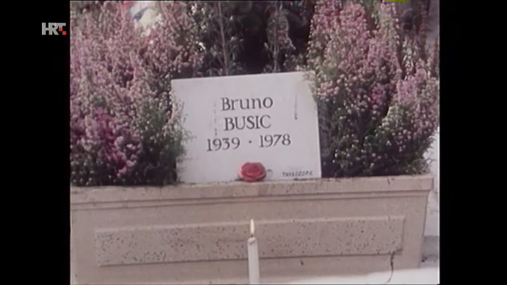 A film frame of a white headstone cut with the name Bruno Busic and the dates 1939 and 1978, banked with flowering heather, a single red rose laid against it and a candle burning below
