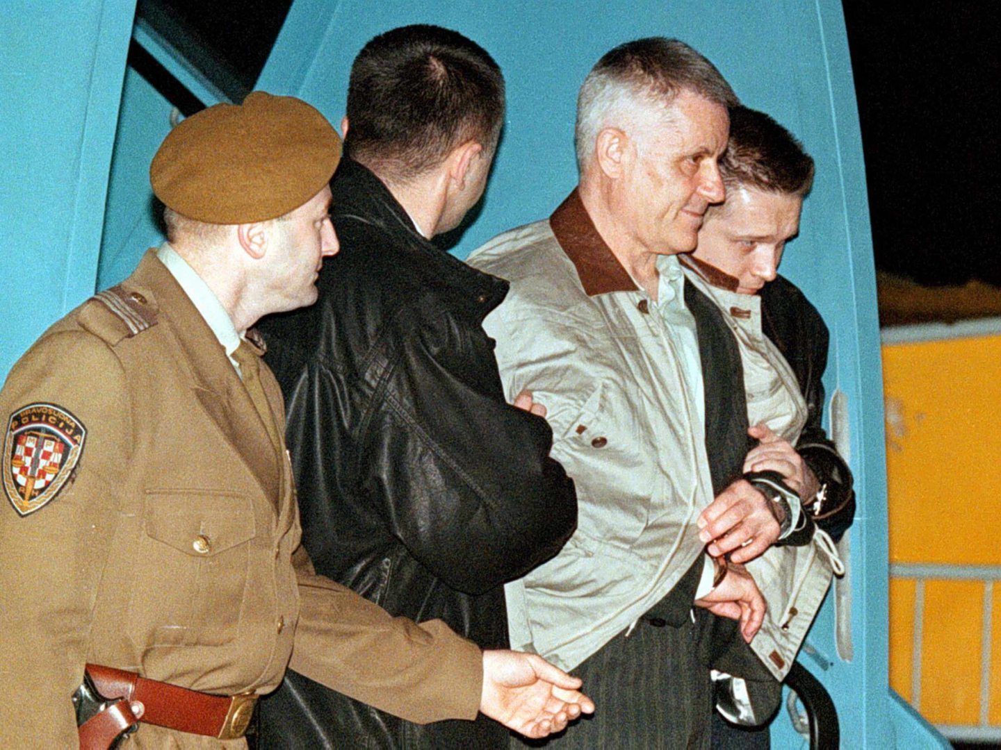 A night photograph of a man in a tan jacket being walked down a set of aircraft steps by escorting officers on either side