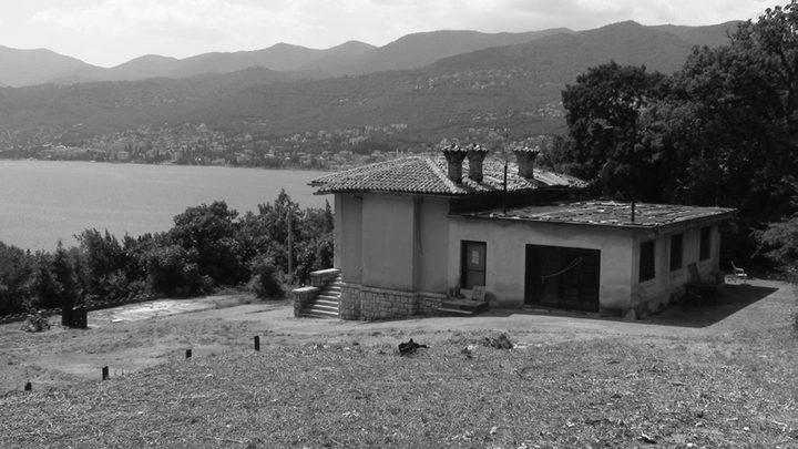 A black and white photograph of a single storey villa with a tiled roof and tall chimneys standing on open ground above the sea, the wooded slopes and scattered houses of the Kvarner coast behind it