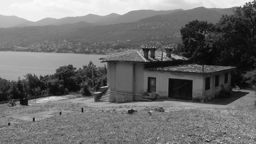 A black and white photograph of a single storey villa with a tiled roof and tall chimneys standing on open ground above the sea, the wooded slopes and scattered houses of the Kvarner coast behind it