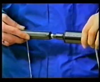 A television frame of two hands screwing a long cylindrical silencer onto the muzzle of a small automatic pistol, against a blue backdrop