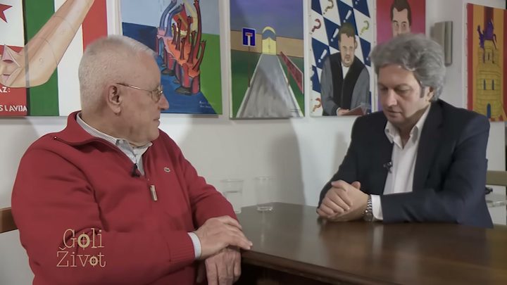 A television frame of an elderly Vinko Sindicic in a red zipped sweater and glasses, sitting at a table opposite his interviewer in a room hung with painted panels