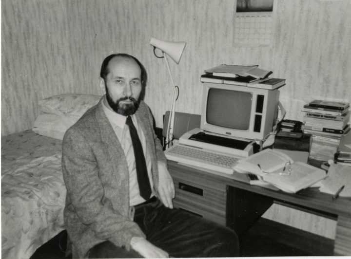 A black and white photograph of Nikola Stedul, bearded and in a jacket and tie, sitting on the edge of a bed beside a desk with an early home computer, an anglepoise lamp, papers and a stack of books