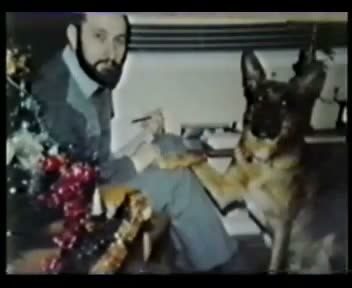 A television frame of a family photograph: a bearded Nikola Stedul sitting at home beside a decorated Christmas tree, holding out a hand to a German shepherd dog at his side