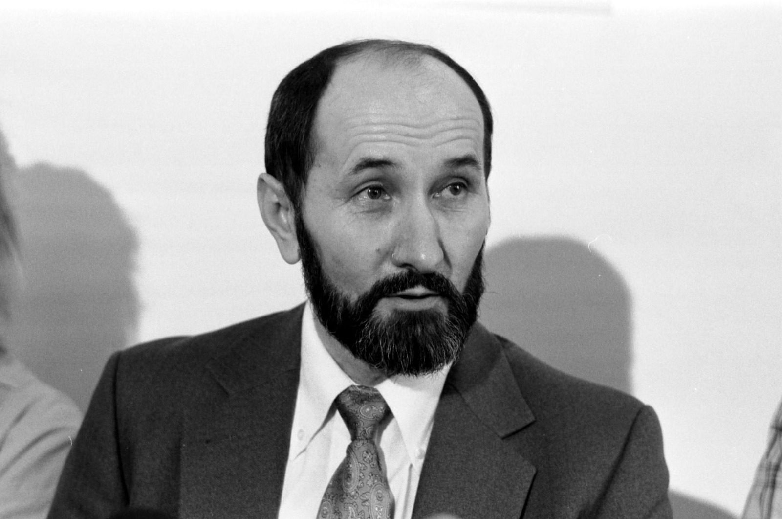 A black and white photograph of Nikola Stedul, bearded and in a suit and patterned tie, speaking at a press conference against a plain wall