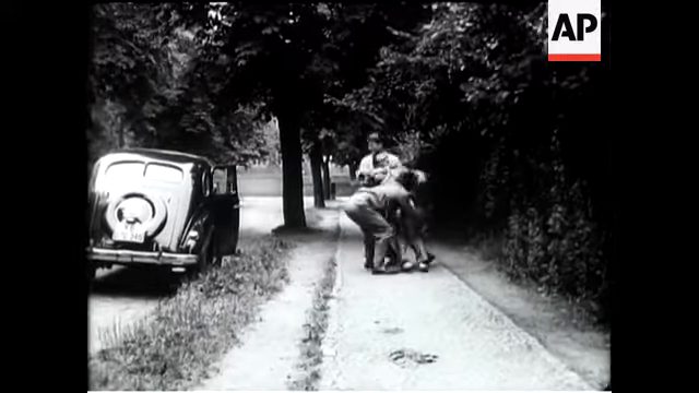 A staged newsreel frame of a man being seized beside a dark saloon on a leafy street, filmed as a reconstruction for a British newsreel in July 1952