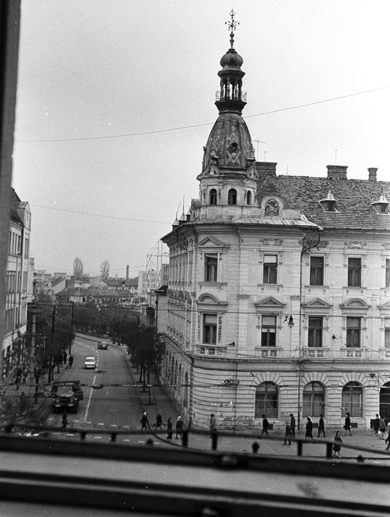 A black and white photograph taken from an upper terrace in Cluj, looking down on a street corner: a nineteenth century corner building with an onion domed tower, a street sign reading str. ZALAU, period cars on the road and people crossing below