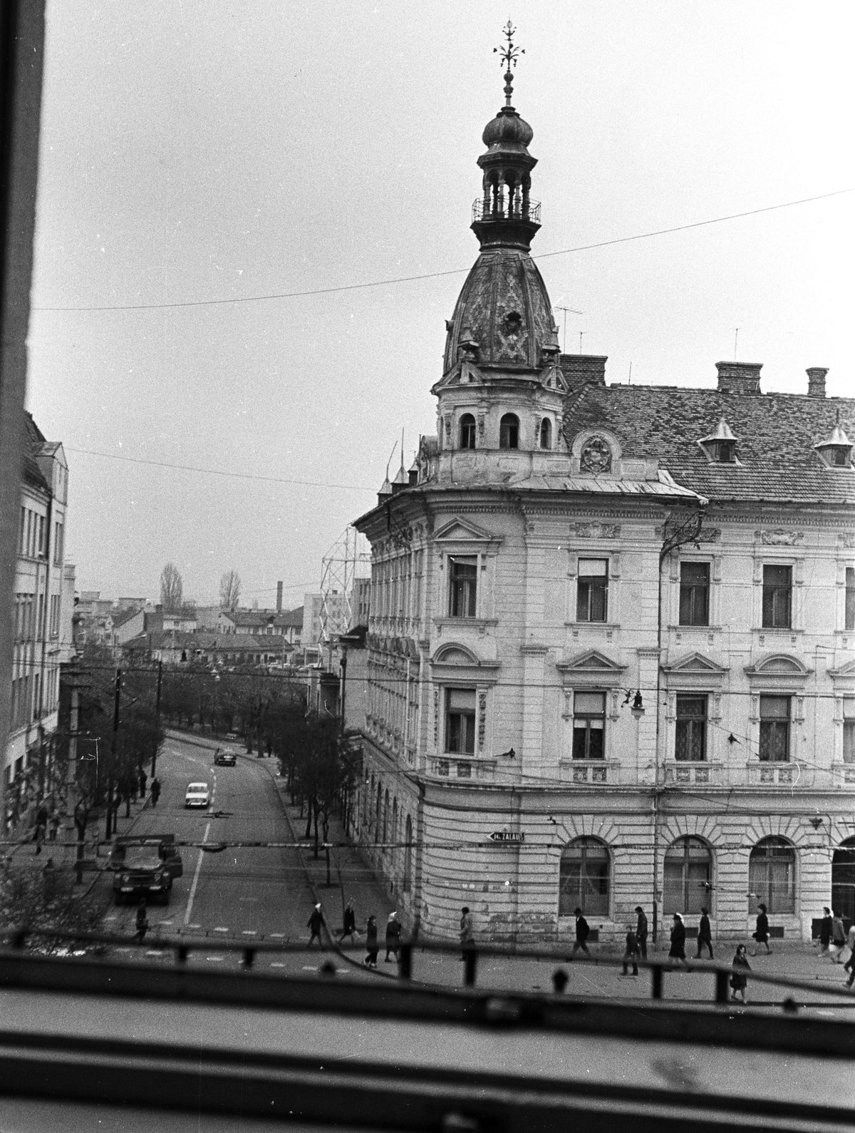 A black and white photograph taken from an upper terrace in Cluj, looking down on a street corner: a nineteenth century corner building with an onion domed tower, a street sign reading str. ZALAU, period cars on the road and people crossing below