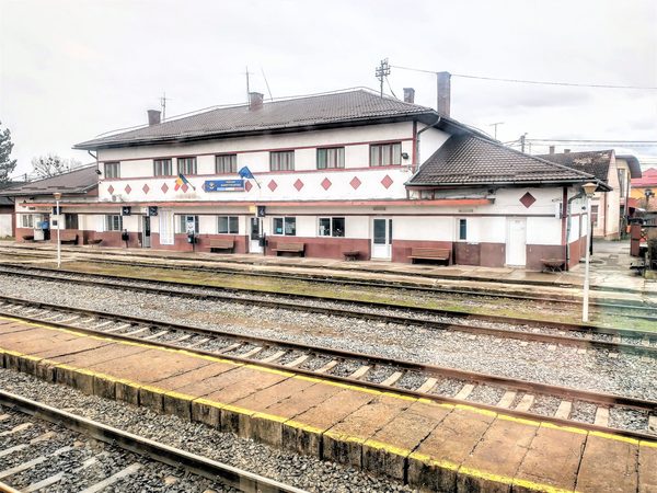 A colour photograph of the platform side of Huedin railway station, a long white building with dark red trim and diamond decoration, carrying a blue sign that reads HUEDIN BANFFYHUNYAD, with benches under the canopy and three tracks in the foreground