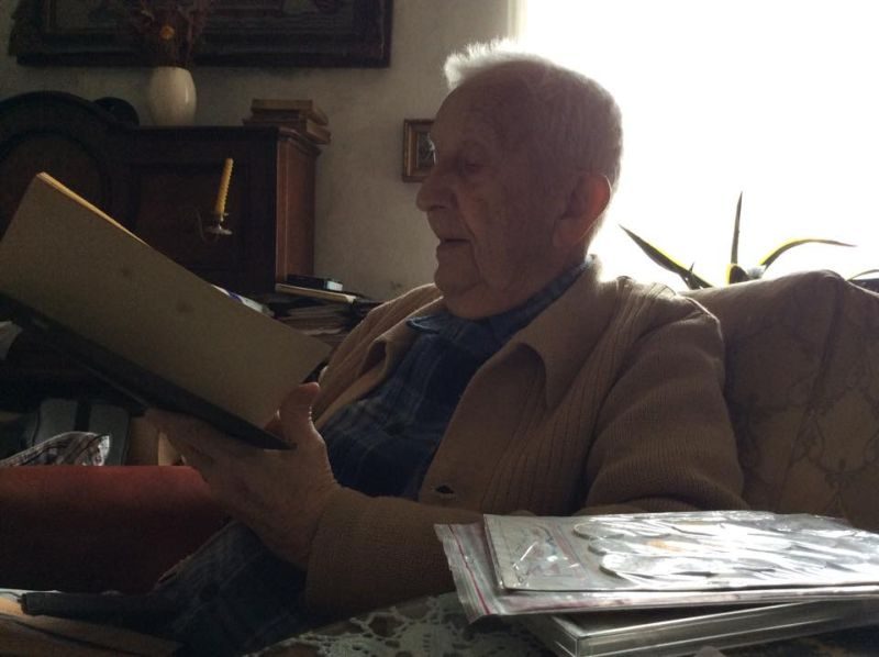 A colour photograph of an elderly man in a cardigan sitting in an armchair at home, holding an open folder and reading aloud from it, with albums and papers on the table beside him