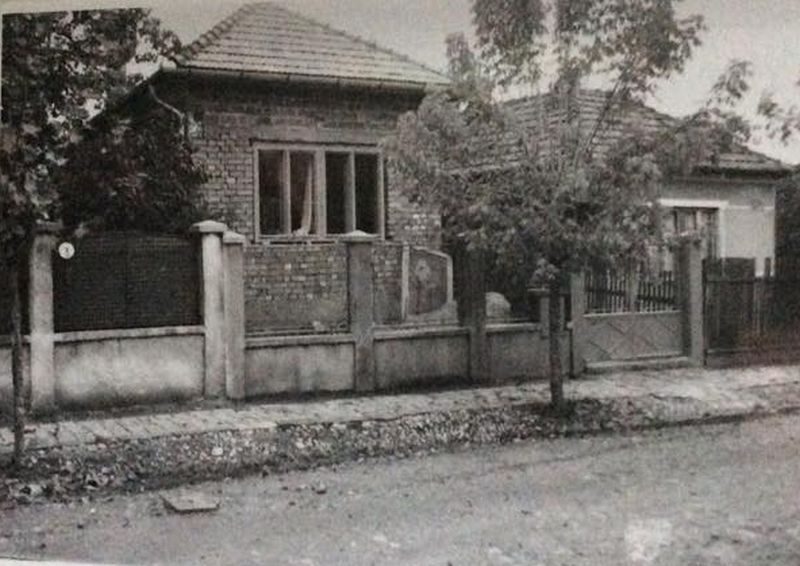 A black and white photograph of a single storey brick house with a tiled roof behind a low concrete fence and a wooden gate, trees in front of it and an unpaved street in the foreground