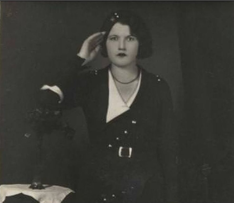 A young woman in a dark belted dress standing beside a table with flowers, in a studio portrait of the early 1930s