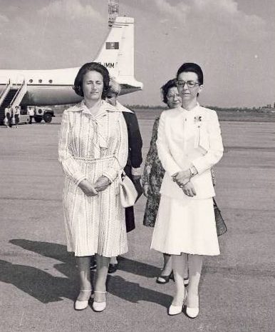 Two women standing on an airfield apron in front of an aircraft, one in a patterned summer dress, the other in a light suit and glasses