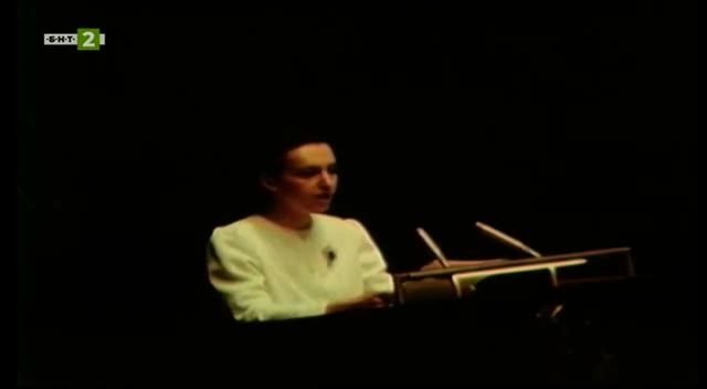 A woman in a white dress speaking at a lectern with two microphones, lit against a dark hall