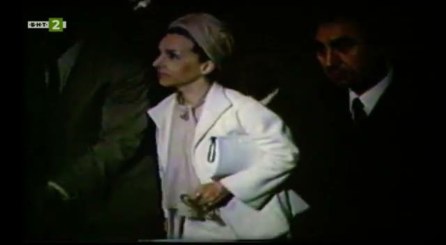Film of a woman in a white coat and a white head wrap standing in a dark interior, a man in a dark suit behind her shoulder