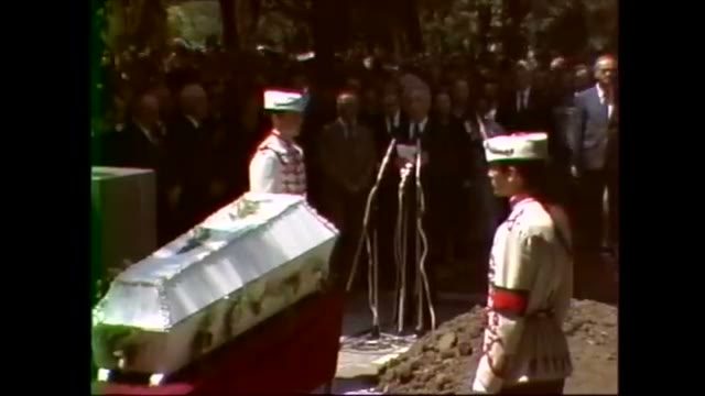 Colour film of a coffin standing among flowers with two guards in white ceremonial tunics and plumed caps on either side, mourners and microphones behind, fresh earth in the foreground