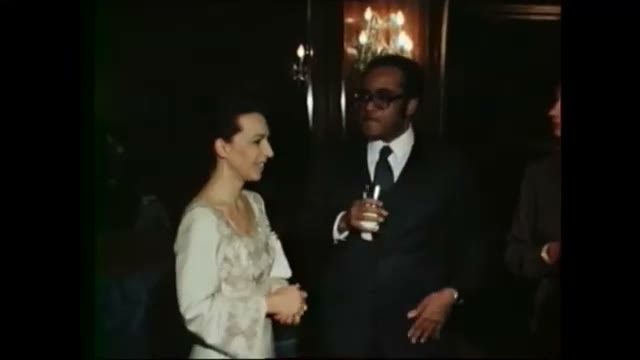 Colour film of a woman in a pale embroidered dress talking with a man in a dark suit at a reception, a chandelier and panelled walls behind them