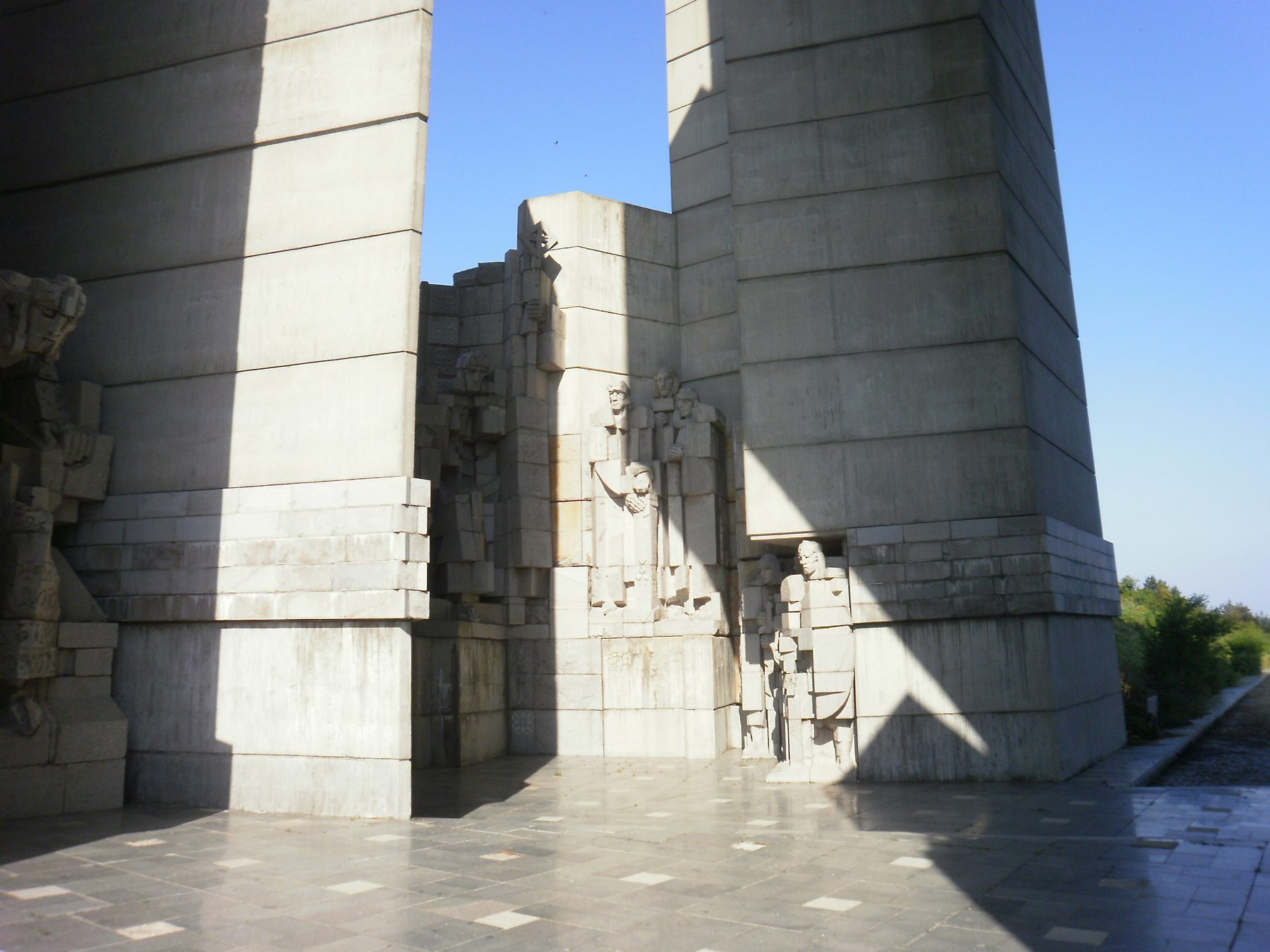 Massive angular concrete blocks carrying carved figures, seen from a paved terrace against a clear sky