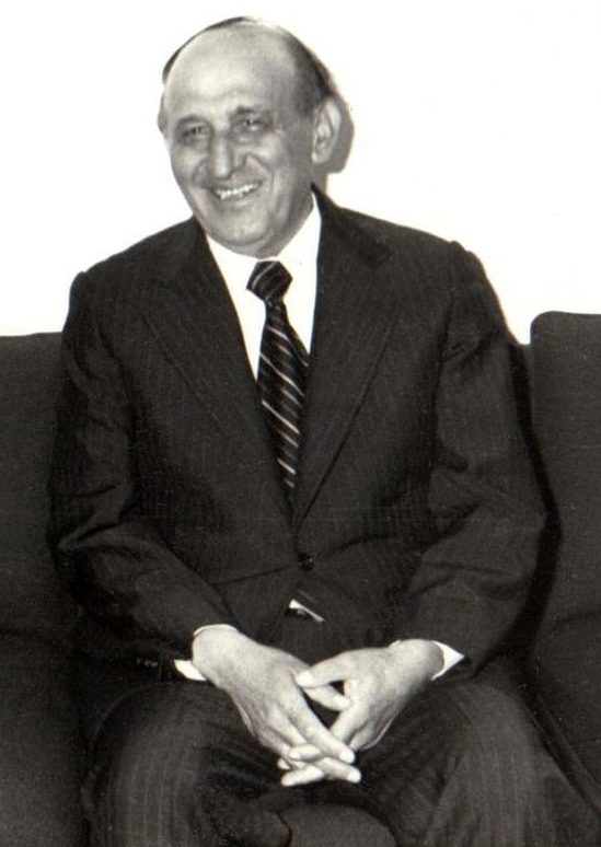 A black and white photograph of a smiling middle aged man in a dark suit and tie, seated with his hands folded