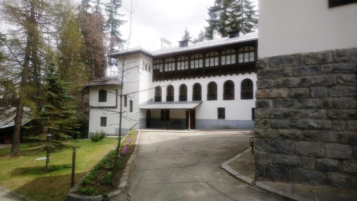 A white mountain residence of two storeys with a long glazed timber gallery above and arched windows below, standing among conifers