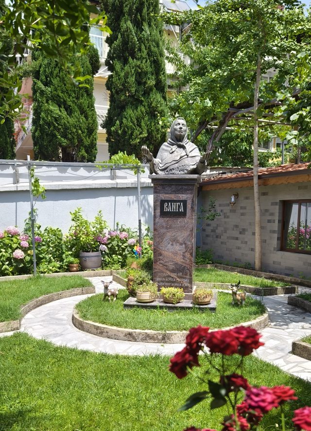 A bronze bust of an elderly woman in a headscarf with her hands raised, on a granite plinth cut with her name in Cyrillic, standing in a garden