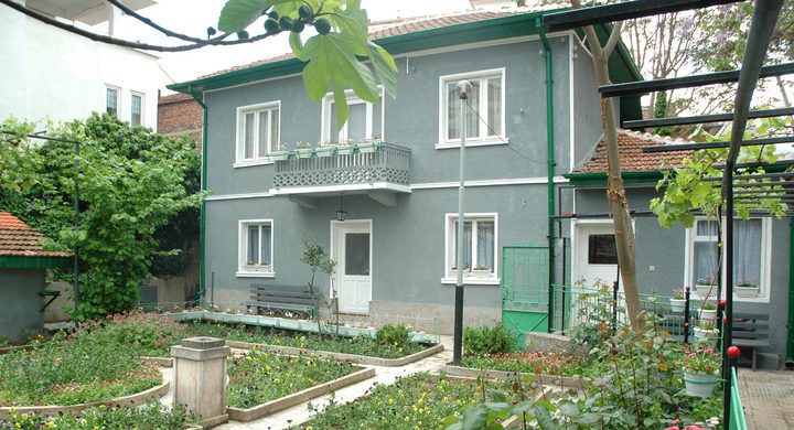 A grey two storey house with white window frames and a small balcony, behind a walled town garden of flower beds and vines